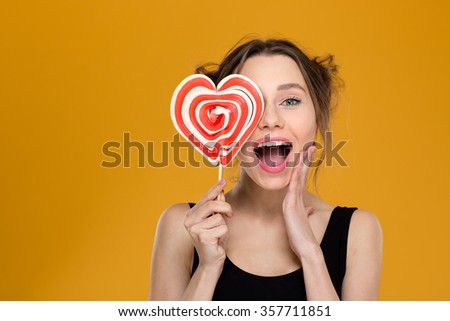 Happy excited young woman covered her eye with bright heart shaped lollipop over yellow background