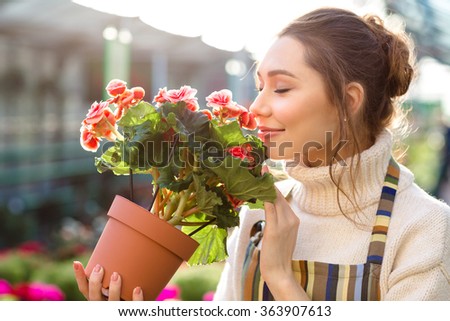 Inspired smiling young woman florist smelling flowers of begonia in greenhouse