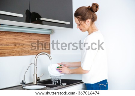 Cute lovely young woman standing and washing dishes at home