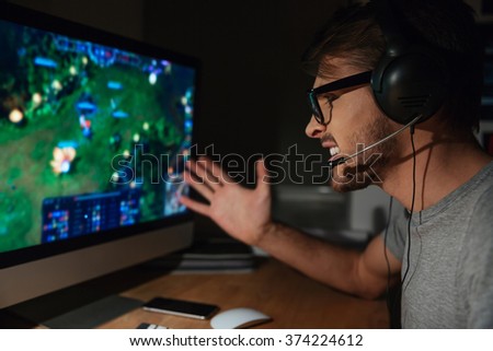 Angry young gamer in glasses playing game on computer using headphones