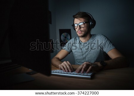 Concentrated young man in headphones sitting in dark room and using computer