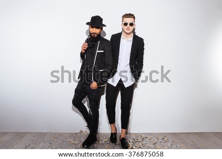 Two attractive confident young men in modern black suits standing over white background
