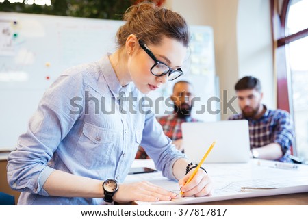 Woman engineer working on blueprint in office with colleagues on background