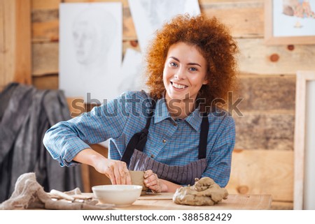 Cheerful young woman potter with curly red hair in apron sitting and working in art pottery studio