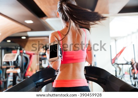 Back view of pretty young woman athlete with balnk screen smartphone running on treadmill in gym