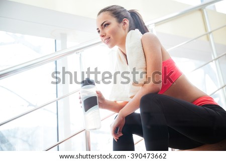 Happy beautiful young sportswoman resting and drinking water on stairs in gym