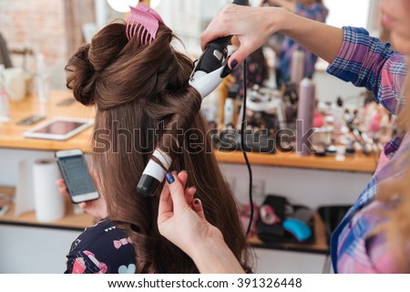 Woman hairdresser making hairstyle using curling iron for long hair of young female with smartphone in beauty salon