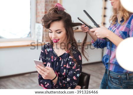 Female hairdresser applying hair straightener for long hair of smiling young woman using smartphone in dressing room
