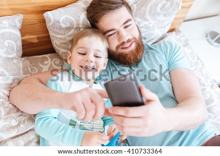 Top view of cheerful bearded father using smartphone and lying on bed with little son