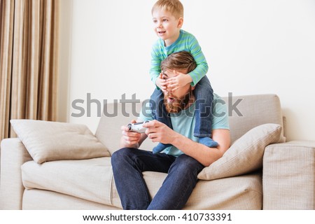 Smiling little boy covering eyes of his father while he playing computer games on sofa at home