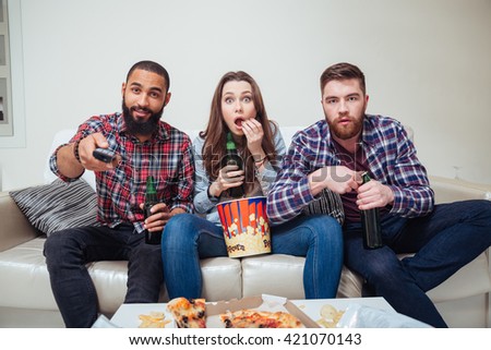 Group of amazed shocked young friends watching tv and eating popcorn on sofa