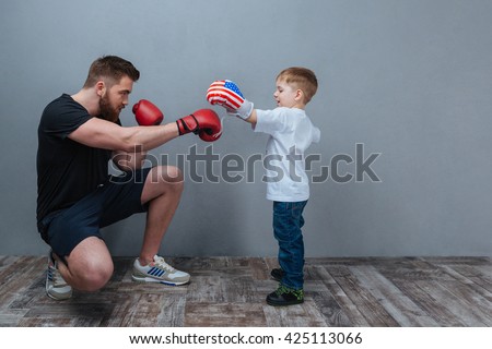 Father and little son working out in boxing gloves together over grey background