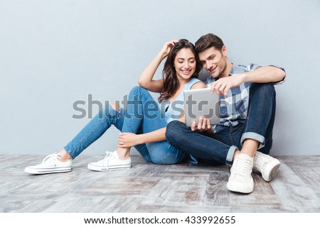 Happy young couple using tablet sitting on the floor at home isolated on gray background