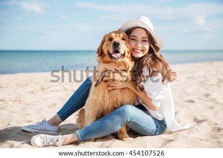 Cheerful pretty young woman in hat sitting and hugging her dog on the beach