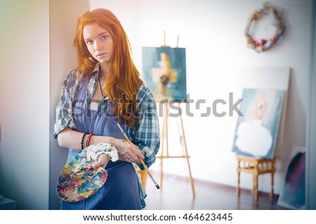 Thoughtful attractive young woman painter with long red hair in apron holding art palette and brush in artist workshop