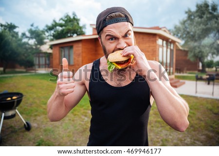 Young bearded hipster guy biting delicious burger and showing thumbs up outdoors