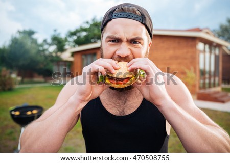 Young bearded hipster guy eating delicious burger outdoors