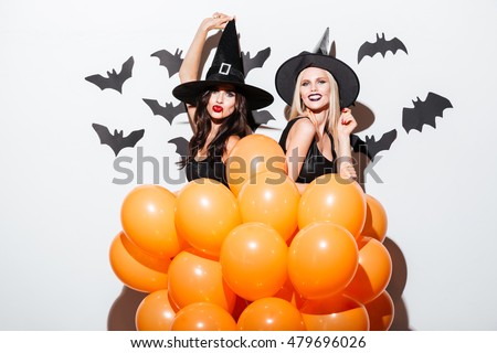 Two cheerful young women in witch halloween costumes dancing with orange balloons over white background