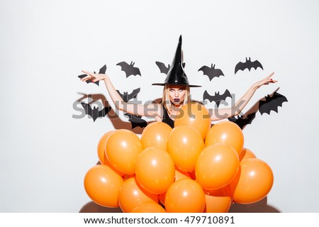 Gothic young woman in witch hat standing with raised hands behind orange balloons over white background