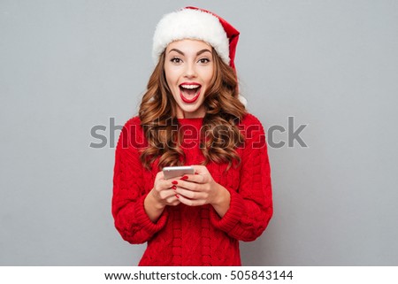 Surprised pretty young woman in santa claus hat using smartphone over gray background