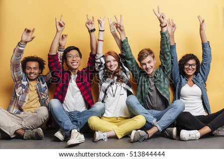 Multiethnic group of smiling young people celebrating success and showing peace sign with raised hands over yellow background