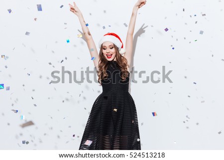 Happy charming young woman in santa claus hat standing with raised hands over white background