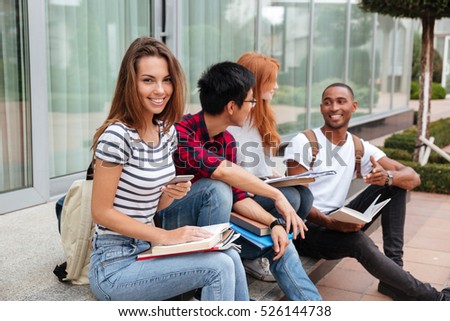 Smiling beautiful young woman sitting with her friends and using mobile phone outdoors
