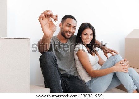 Loving couple sitting with unpacked boxes. Man holding key and showing it to camera. Looking at camera. Focus on key.