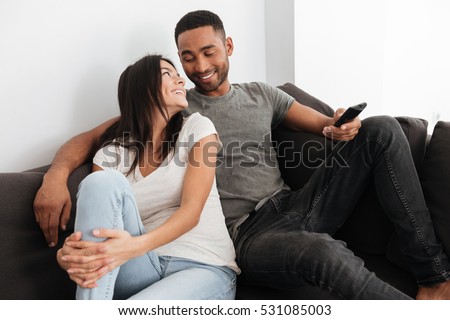 Image of happy cute young couple hugging and watching TV on sofa at home. Look at each other.