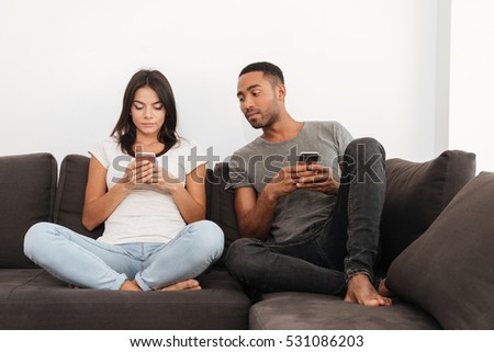 Photo of young couple using their cellphones sitting on sofa at home. Boy looking at woman\'s phone.