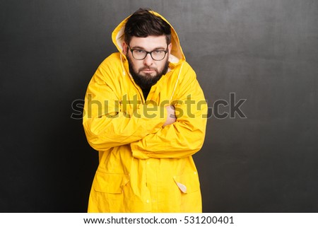 Portrait of bearded man dressed in raincoat and wearing glasses standing over chalkboard with arms crossed. Look at camera.