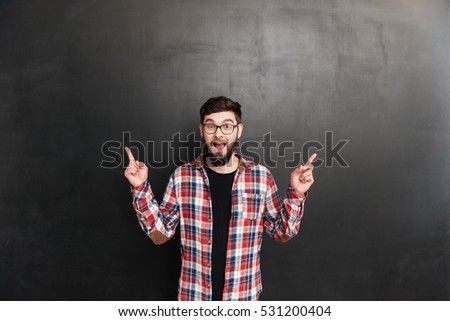 Happy bearded young man in glasses pointing away with both hands over grey background