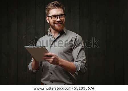 Portrait of a smiling happy man in eyeglasses holding tablet computer and looking away isolated on the black wooden background