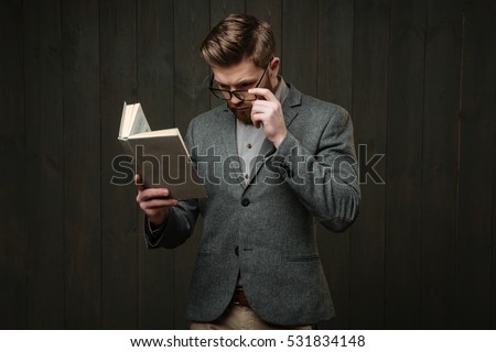 Portrait of a thoughtful bearded man in eyeglasses and jacket reading book isolated on the black wooden background