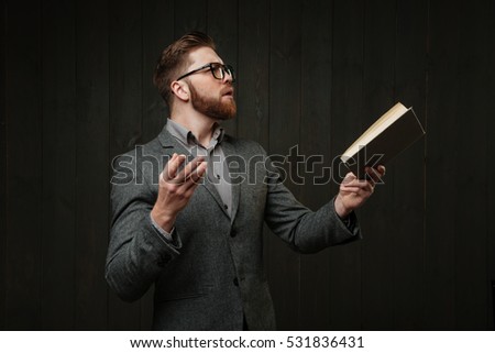 Bearded young man in eyeglasses and casual suit reading book and gesturing with hands isolated on the black wooden background