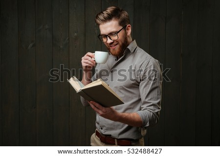 Portrait of a smiling bearded man in eyeglasses reading book and drinking cup of coffee isolated on a black wooden background