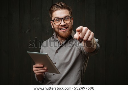 Close up portrait of a smiling happy man in eyeglasses holding tablet computer and pointing at camera isolated on a black wooden background