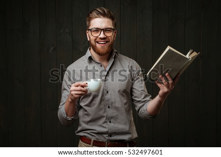 Portrait of a cheerful happy man in eyeglasses and shirt reading book and holding cup of coffee isolated on a black wooden background