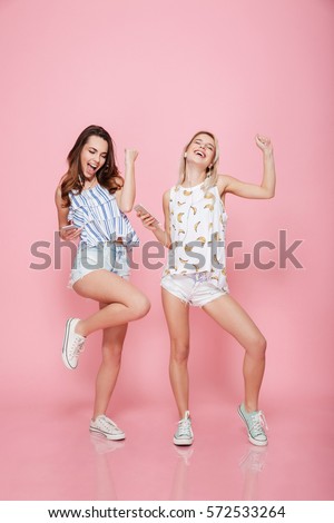 Full length portrait of a happy girlfriends dancing and having fun while listening to music isolated on the pink background