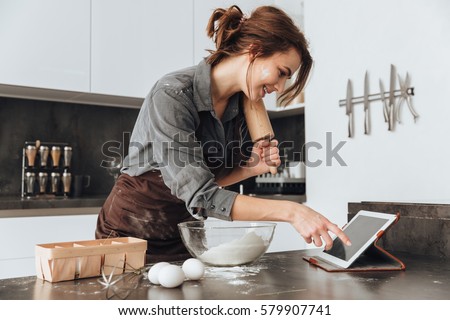 Image of young pretty lady standing in kitchen and cooking the dough. Looking at tablet computer.