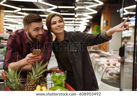 Image of young happy loving couple in supermarket with shopping trolley choosing products. Man holding mobile phone.