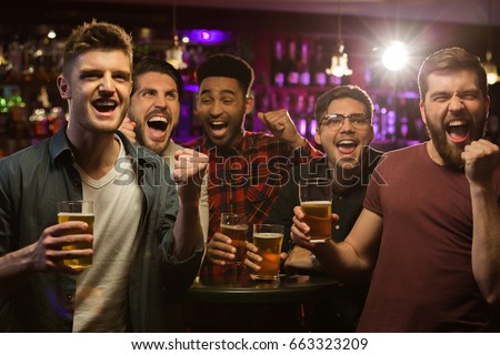 Four happy men holding beer mugs and gesturing while watching TV in bar or pub