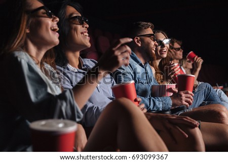 Image of young happy friends sitting in cinema watch film eating popcorn.