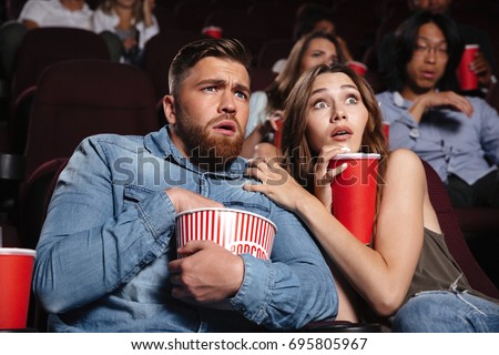 Young scared couple watching a horror movie while sitting at the cinema with popcorn