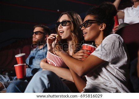 Two young women in eyeglasses watching 3d movie and eating popcorn while sitting in a cinema
