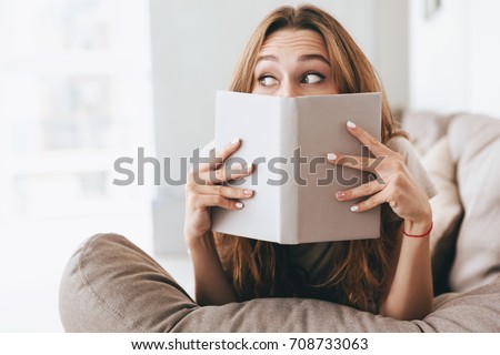 Photo of young pretty lady sitting on sofa indoors. Looking aside covering face with book.