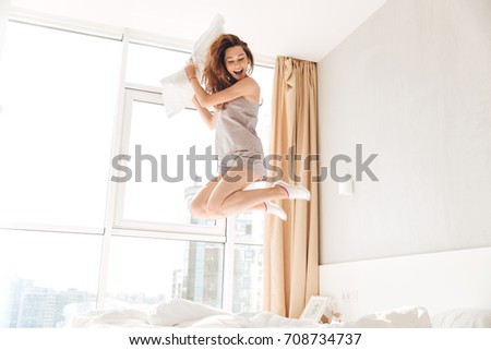 Picture of young happy pretty lady jumping on bed holding pillow indoors. Looking aside.
