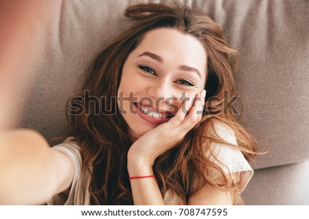 Picture of young amazing happy emotional pretty lady lies on sofa indoors. Looking camera make selfie.