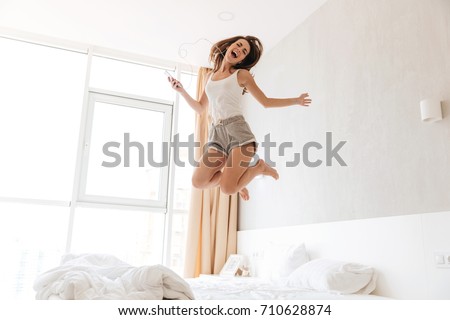 Full length portrait of a cheerful singing woman in earphones listening to music and jumping on bed indoors