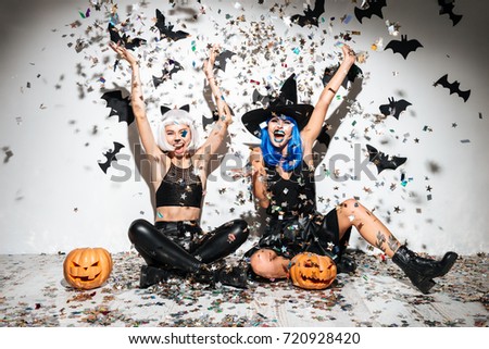 Two funny young women in leather halloween costumes posing with curved pumpkins over bats and confetti background
